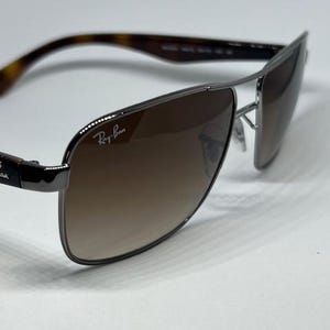 Pode incluir: &Oacute;culos de sol Ray-Ban com arma&ccedil;&atilde;o de metal cinza chumbo e design de ponte dupla. As lentes s&atilde;o castanhas degrad&ecirc; e a lente direita exibe o log&oacute;tipo branco da Ray-Ban. As hastes t&ecirc;m um padr&atilde;o de tartaruga em tons de castanho e preto.