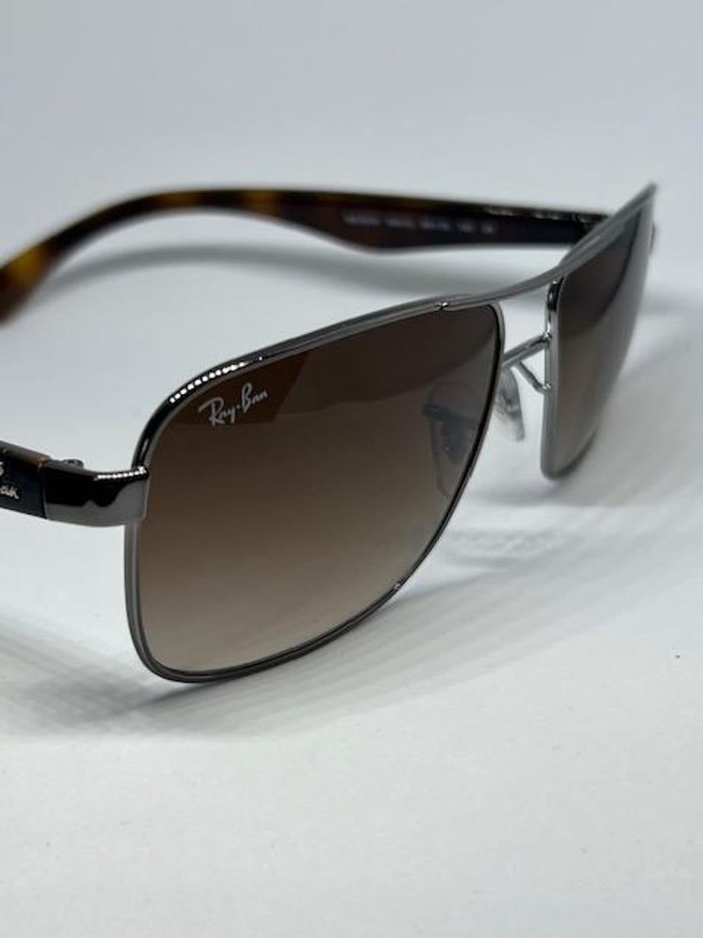 Pode incluir: &Oacute;culos de sol Ray-Ban com arma&ccedil;&atilde;o de metal cinza chumbo e design de ponte dupla. As lentes s&atilde;o castanhas degrad&ecirc; e a lente direita exibe o log&oacute;tipo branco da Ray-Ban. As hastes t&ecirc;m um padr&atilde;o de tartaruga em tons de castanho e preto.