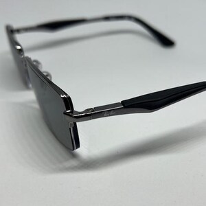 Puede incluir: Gafas de sol rectangulares con montura de metal gris oscuro y puntas de patilla negras. Las lentes son oscuras y reflectantes. El logotipo de Ray-Ban es visible en la bisagra met&aacute;lica de la patilla. Dise&ntilde;o con puente fino y brazos delgados.