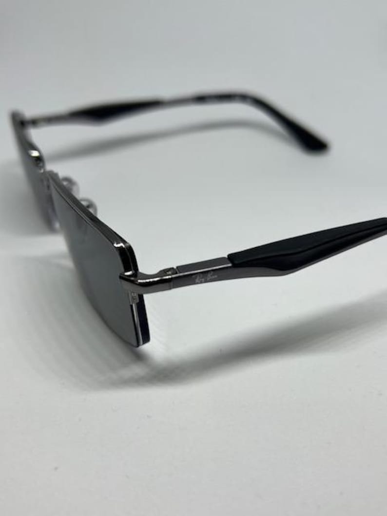 Puede incluir: Gafas de sol rectangulares con montura de metal gris oscuro y puntas de patilla negras. Las lentes son oscuras y reflectantes. El logotipo de Ray-Ban es visible en la bisagra met&aacute;lica de la patilla. Dise&ntilde;o con puente fino y brazos delgados.