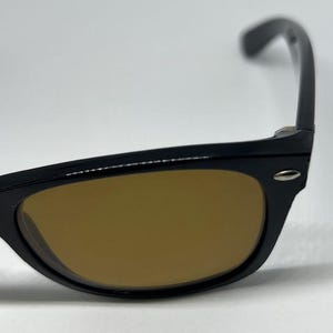 Puede incluir: Gafas de sol con montura negra y lentes tintadas en color &aacute;mbar. La montura tiene una forma cl&aacute;sica con un peque&ntilde;o detalle met&aacute;lico plateado en la bisagra donde la patilla se une al borde frontal. Las patillas son curvas y se extienden hacia atr&aacute;s.