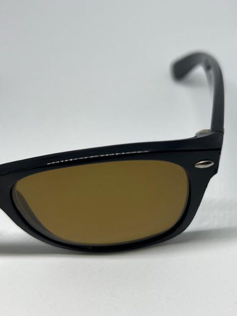 Puede incluir: Gafas de sol con montura negra y lentes tintadas en color &aacute;mbar. La montura tiene una forma cl&aacute;sica con un peque&ntilde;o detalle met&aacute;lico plateado en la bisagra donde la patilla se une al borde frontal. Las patillas son curvas y se extienden hacia atr&aacute;s.