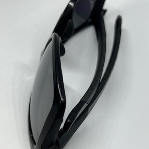 May include: A pair of black sunglasses with dark, tinted lenses and a matte black frame. The sunglasses are shown from a side angle, displaying the curved temples and the structure of the frame. The design features a solid black finish throughout.
