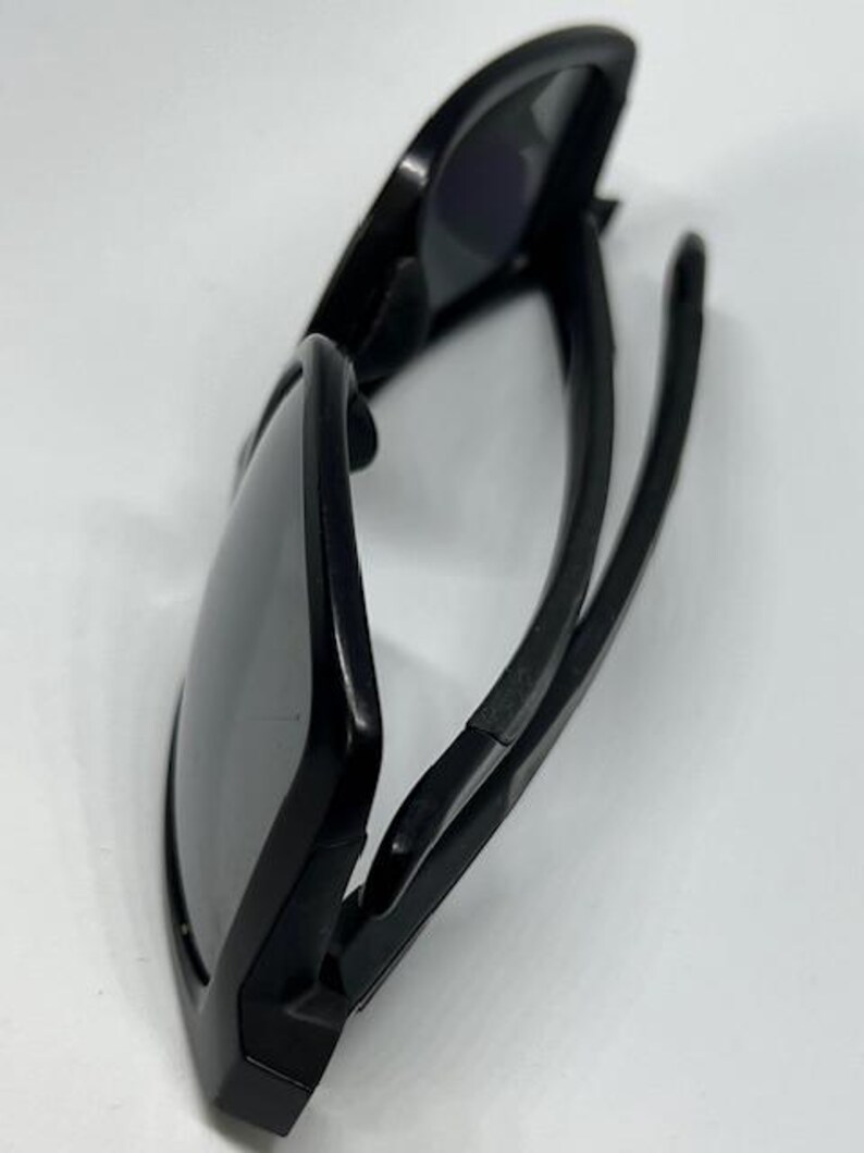 May include: A pair of black sunglasses with dark, tinted lenses and a matte black frame. The sunglasses are shown from a side angle, displaying the curved temples and the structure of the frame. The design features a solid black finish throughout.