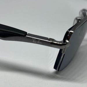 Puede incluir: Unas gafas de sol con montura met&aacute;lica oscura y patillas negras. El lateral de la patilla met&aacute;lica lleva grabado el logotipo blanco de Ray-Ban. Las lentes son oscuras y de forma rectangular, unidas a la patilla mediante una peque&ntilde;a bisagra.