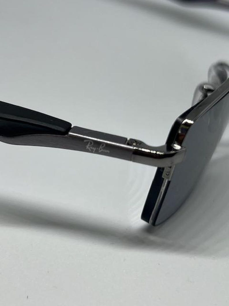Puede incluir: Unas gafas de sol con montura met&aacute;lica oscura y patillas negras. El lateral de la patilla met&aacute;lica lleva grabado el logotipo blanco de Ray-Ban. Las lentes son oscuras y de forma rectangular, unidas a la patilla mediante una peque&ntilde;a bisagra.