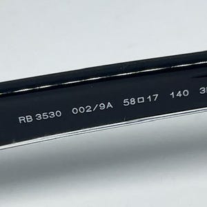 May include: The interior of a black eyeglass temple arm featuring white printed text that reads RB 3530 002/9A 58 17 140 3P. The surface is smooth and glossy, showing the model number, color code, lens width, bridge width, and temple length measurements.