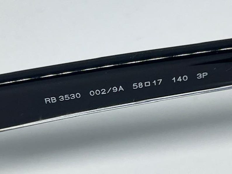 May include: The interior of a black eyeglass temple arm featuring white printed text that reads RB 3530 002/9A 58 17 140 3P. The surface is smooth and glossy, showing the model number, color code, lens width, bridge width, and temple length measurements.
