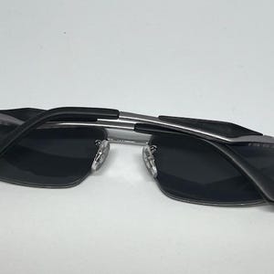 Puede incluir: Gafas de sol rectangulares con lentes gris oscuro y montura de metal plateado. Las patillas son negras con un detalle plateado, y el logotipo de Ray-Ban es visible en la esquina superior de la lente y en la patilla. El puente tiene almohadillas.
