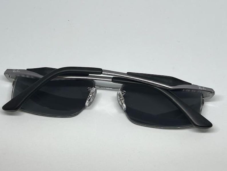 Puede incluir: Gafas de sol rectangulares con lentes gris oscuro y montura de metal plateado. Las patillas son negras con un detalle plateado, y el logotipo de Ray-Ban es visible en la esquina superior de la lente y en la patilla. El puente tiene almohadillas.