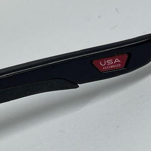 May include: A black temple arm of a pair of glasses featuring a small, rectangular red inlay near the tip. The red inlay contains the white text USA ASSEMBLED. The arm has a smooth, matte black finish with a slightly textured black grip section.