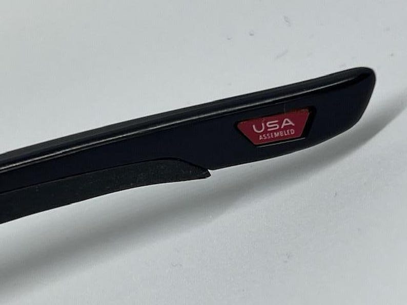 May include: A black temple arm of a pair of glasses featuring a small, rectangular red inlay near the tip. The red inlay contains the white text USA ASSEMBLED. The arm has a smooth, matte black finish with a slightly textured black grip section.