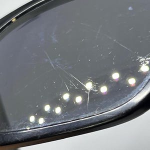 May include: A pair of black-framed sunglasses featuring dark, tinted lenses. The surface of the lens shows visible surface scratches and scuffs, with multiple small, bright light reflections scattered across the dark, reflective glass material.