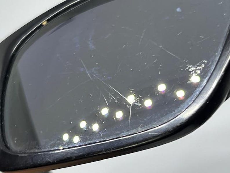 May include: A pair of black-framed sunglasses featuring dark, tinted lenses. The surface of the lens shows visible surface scratches and scuffs, with multiple small, bright light reflections scattered across the dark, reflective glass material.