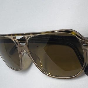 May include: Aviator sunglasses featuring a translucent beige frame with a tortoiseshell pattern along the top brow bar. The lenses are a dark brown tint, and the frame includes a double bridge design with thin, clear plastic nose pads.