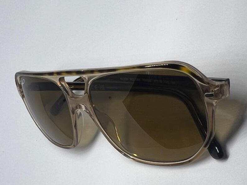 May include: Aviator sunglasses featuring a translucent beige frame with a tortoiseshell pattern along the top brow bar. The lenses are a dark brown tint, and the frame includes a double bridge design with thin, clear plastic nose pads.