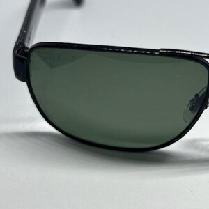 May include: Black-framed sunglasses featuring dark green lenses with a subtle Ray-Ban logo etched into the upper corner of the right lens. The frames have a thin metal bridge and black temples, showcasing a classic design with a polished finish.