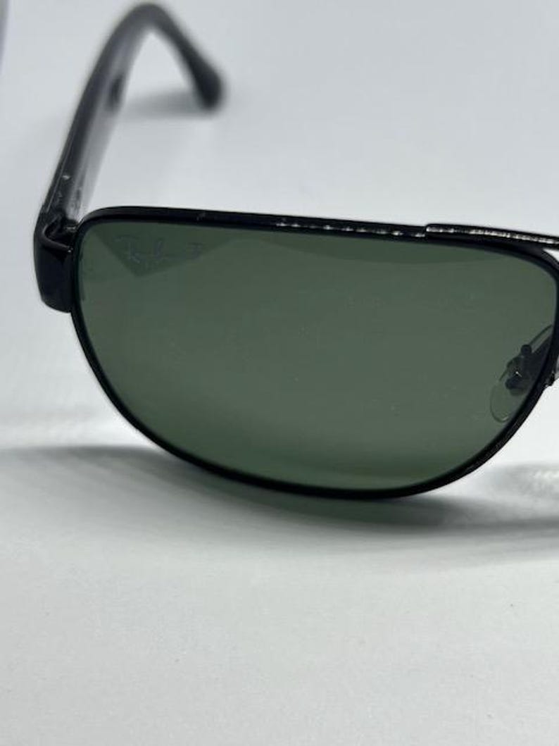 May include: Black-framed sunglasses featuring dark green lenses with a subtle Ray-Ban logo etched into the upper corner of the right lens. The frames have a thin metal bridge and black temples, showcasing a classic design with a polished finish.