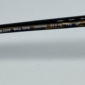 May include: The inner temple arm of a pair of sunglasses featuring a dark brown tortoiseshell pattern. Printed in white text on the arm are the model details: RB 2205 BILL ONE 1292/M2 57 16 145 3P. The arm has a smooth, glossy finish and a metallic core.