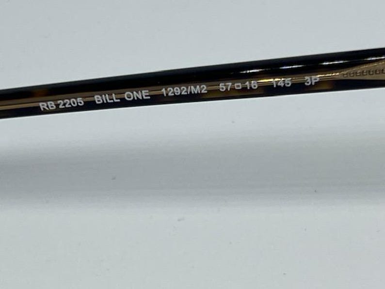 May include: The inner temple arm of a pair of sunglasses featuring a dark brown tortoiseshell pattern. Printed in white text on the arm are the model details: RB 2205 BILL ONE 1292/M2 57 16 145 3P. The arm has a smooth, glossy finish and a metallic core.