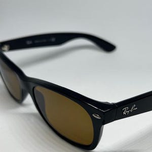 Puede incluir: Gafas de sol negras con lentes tintadas en marr&oacute;n y acabado brillante. La montura tiene una forma rectangular cl&aacute;sica con detalles met&aacute;licos plateados en las bisagras. El logotipo de Ray-Ban es visible en la patilla de la gafa.