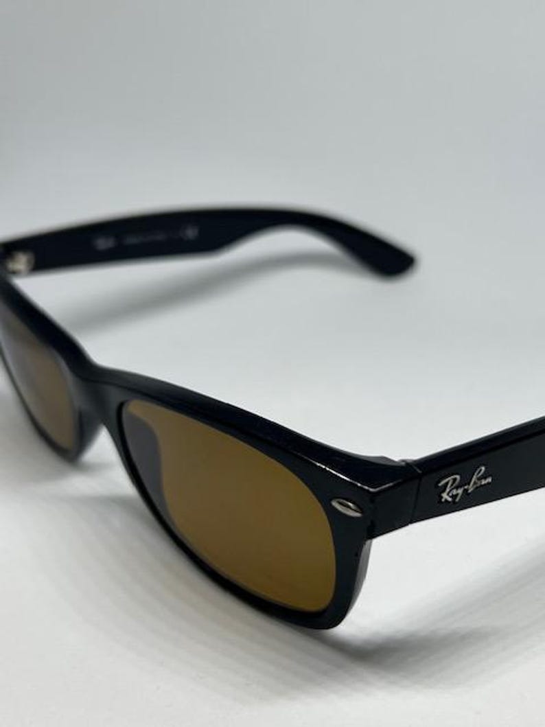 Puede incluir: Gafas de sol negras con lentes tintadas en marr&oacute;n y acabado brillante. La montura tiene una forma rectangular cl&aacute;sica con detalles met&aacute;licos plateados en las bisagras. El logotipo de Ray-Ban es visible en la patilla de la gafa.