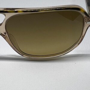 May include: Sunglasses featuring a translucent beige frame with a dark brown tortoiseshell pattern along the top brow bar. The lenses are a solid, opaque brown colour. The frame has a curved, rounded shape with thin, matching beige temples.