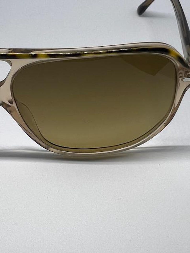 May include: Sunglasses featuring a translucent beige frame with a dark brown tortoiseshell pattern along the top brow bar. The lenses are a solid, opaque brown colour. The frame has a curved, rounded shape with thin, matching beige temples.