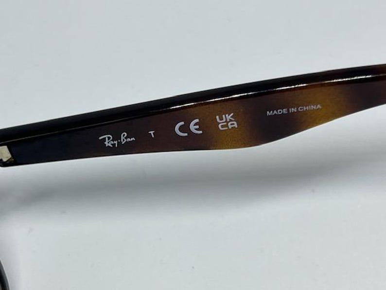 Pode incluir: Uma haste de &oacute;culos com padr&atilde;o de tartaruga castanho e preto, apresentando o texto impresso em branco Ray-Ban, a letra T, a marca CE, a marca UKCA e o texto MADE IN CHINA na superf&iacute;cie interior da haste.