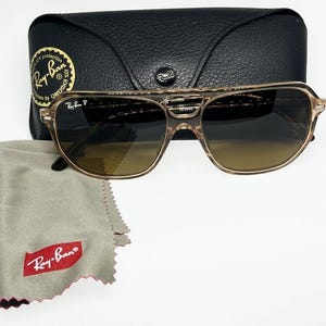 May include: Ray-Ban sunglasses with a translucent tan frame and dark gradient lenses. The glasses feature a double bridge design, the Ray-Ban P logo on the lens, a black leather-textured carrying case with a snap closure, and a grey cleaning cloth.