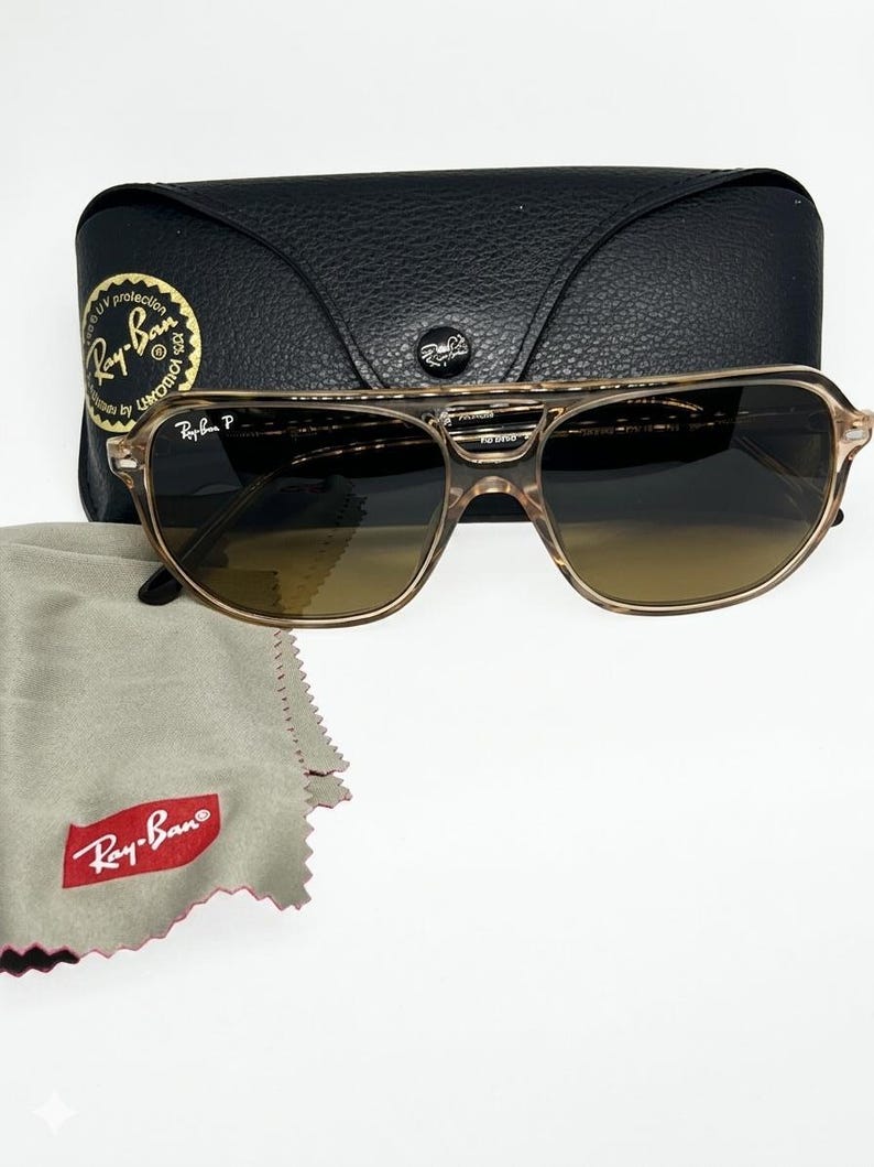 May include: Ray-Ban sunglasses with a translucent tan frame and dark gradient lenses. The glasses feature a double bridge design, the Ray-Ban P logo on the lens, a black leather-textured carrying case with a snap closure, and a grey cleaning cloth.