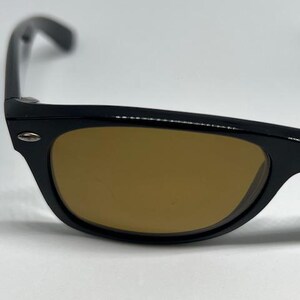 Puede incluir: Un par de gafas de sol con montura de pl&aacute;stico negro brillante y lentes tintadas en marr&oacute;n. La montura presenta una forma rectangular cl&aacute;sica con un peque&ntilde;o detalle de metal plateado en la esquina exterior del borde frontal.