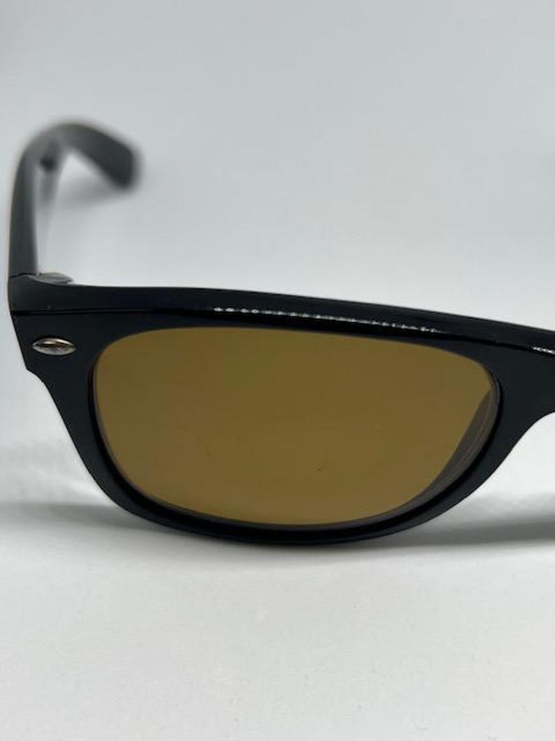 Puede incluir: Un par de gafas de sol con montura de pl&aacute;stico negro brillante y lentes tintadas en marr&oacute;n. La montura presenta una forma rectangular cl&aacute;sica con un peque&ntilde;o detalle de metal plateado en la esquina exterior del borde frontal.