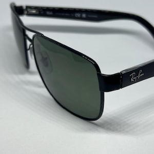 May include: Black Ray-Ban sunglasses featuring a rectangular frame design with dark green tinted lenses. The temples display the silver Ray-Ban logo on the side, and the thin metal bridge connects the two lenses, providing a sleek and structured look.