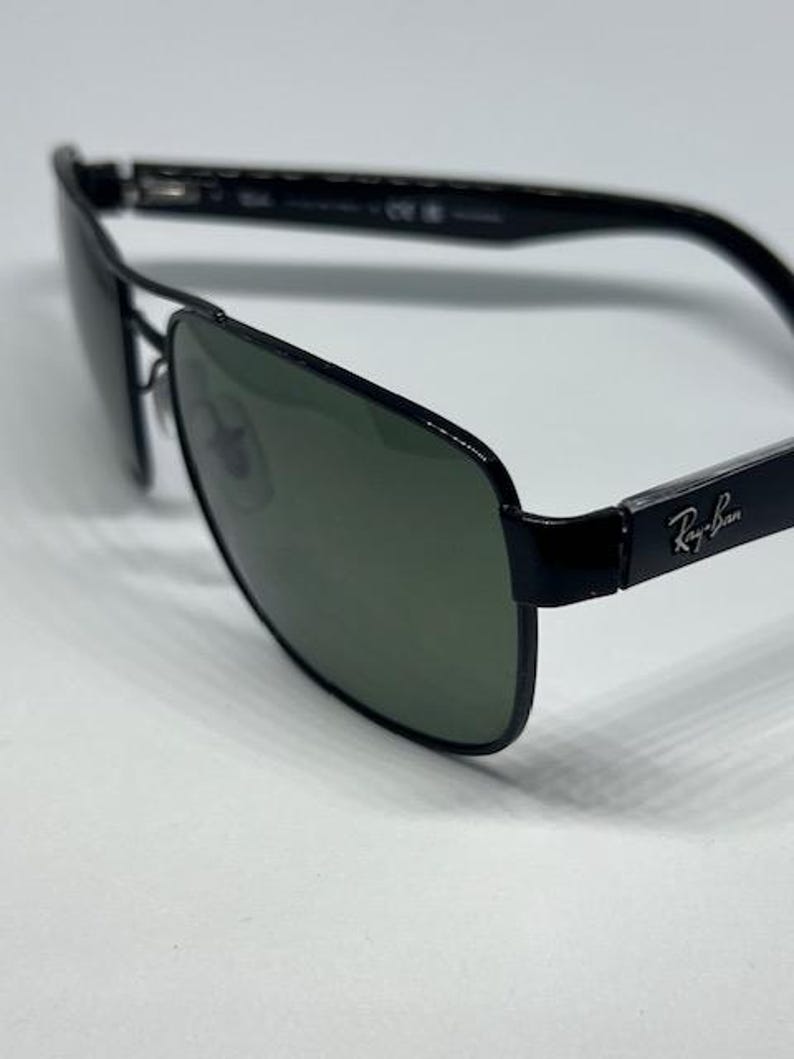 May include: Black Ray-Ban sunglasses featuring a rectangular frame design with dark green tinted lenses. The temples display the silver Ray-Ban logo on the side, and the thin metal bridge connects the two lenses, providing a sleek and structured look.