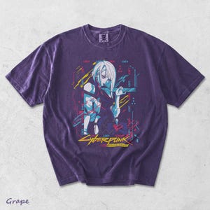 May include: A grape-coloured short-sleeve t-shirt featuring a graphic print of a white-haired character. The design includes the text "LUCY" repeated multiple times, along with "CYBERPUNK" and "EDGERUNNERS" in yellow and red lettering.