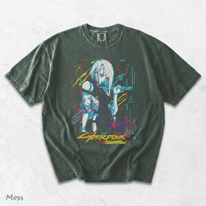 May include: A dark moss green short-sleeve t-shirt featuring a graphic print of a character with white hair. The design includes neon blue, pink, and yellow accents with the text "LUCY" repeated and "CYBERPUNK EDGERUNNERS" printed at the bottom.