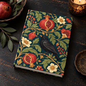 May include: A hardcover journal featuring a dark teal background with a repeating pattern of red pomegranates, cream-colored flowers, green foliage, and a central black bird perched on a branch. The book has a rectangular shape with white pages.