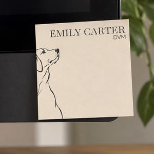 May include: A square beige sticky note featuring the black printed text EMILY CARTER DVM at the top and a black line art illustration of a dog profile on the left side. The note is attached to a black surface.