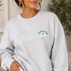 May include: A pale grey sweatshirt featuring the words "rack it club" in green script and a small graphic of three billiard balls. The sweatshirt has a crew neck and is made of a soft material.