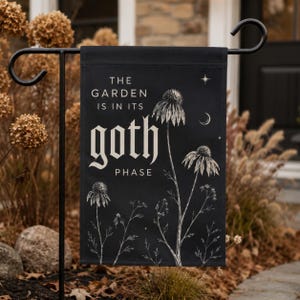 Könnte beinhalten: Eine schwarze, rechteckige Gartenflagge mit weißen botanischen Illustrationen von Sonnenhüten und zarten Wildblumen. Der Text THE GARDEN IS IN ITS goth PHASE ist in einer Mischung aus Serifenschrift und gotischem Stil gehalten.
