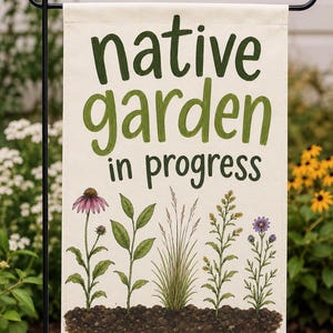 May include: A rectangular off-white garden flag featuring the dark green text "native garden in progress" above an illustration of five distinct plants growing from a patch of brown soil, including a purple coneflower and yellow wildflowers.
