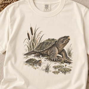 May include: An off-white short-sleeve t-shirt featuring a detailed graphic of a snapping turtle in a marshy environment with cattails and lily pads. The neck label reads Comfort Colors, size L, made in Portugal. The design shows a large turtle.
