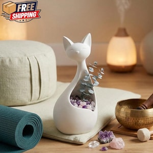 May include: A white ceramic cat-shaped planter filled with purple and clear crystals and sprigs of greenery. A rolled teal yoga mat, a meditation cushion, and a brass singing bowl are also visible.