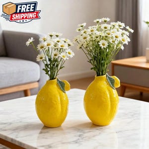 May include: Two bright yellow ceramic vases shaped like lemons, each holding a bouquet of white daisies. The vases have a textured surface and a green leaf detail. The vases are on a marble table.