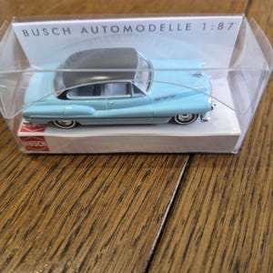 Op de afbeelding: Een lichtblauw vintage automodel met een zwart dak, gepresenteerd in een doorzichtige plastic doos. Het model is een replica op schaal 1:87. De verpakking bevat de merknaam "BUSCH AUTOMODELLE".