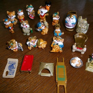 May include: A collection of small, painted plastic cat figurines and miniature accessories. The set includes various anthropomorphic cats in Egyptian-themed attire, a small throne, a red bed, a pyramid structure, a scroll with hieroglyphics, and food.