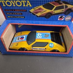Pode incluir: Um carro de corrida de brinquedo amarelo com faixas de corrida azuis e vermelhas, o número 48 e o texto Paris-Dakar, Elf e Turbo impresso na carroceria. O carro está alojado em uma caixa azul com um padrão de grade e um gráfico de um Toyota.