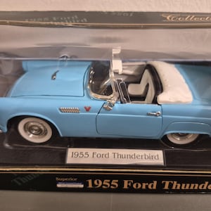 Könnte beinhalten: Ein hellblaues 1955 Ford Thunderbird Modellauto mit weißem Innenraum und weißem Verdeck, präsentiert in einer schwarzen Schachtel mit dem Text 1955 Ford Thunderbird auf dem vorderen Etikett und der Verpackung.