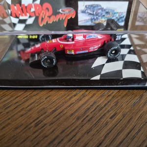 Könnte beinhalten: Rotes Ferrari Formel-1-Rennwagenmodell in einer transparenten Vitrine. Das Modell hat schwarze Reifen und die Nummer 28 auf der Seite. Die Vitrine steht auf einer schwarz-weißen Schachbrettbasis.